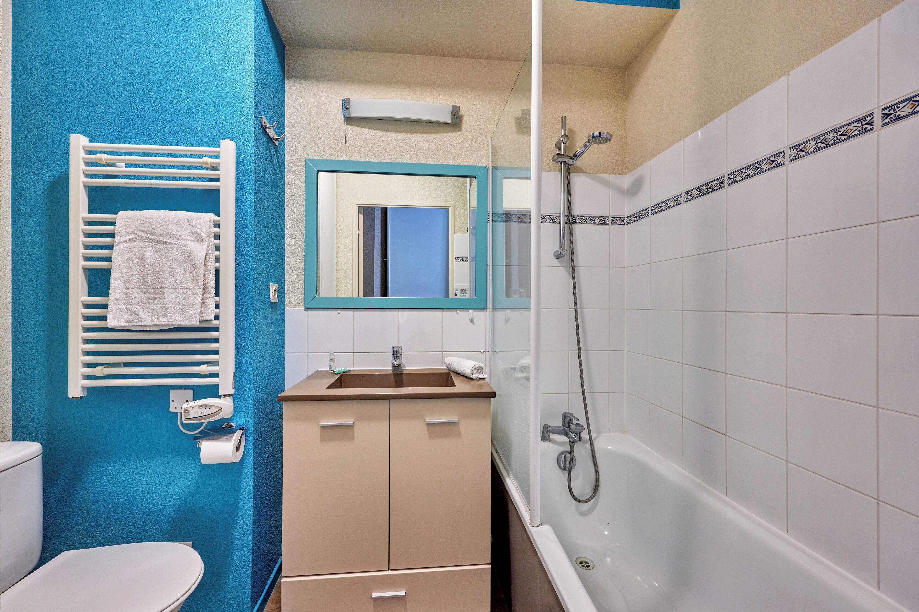 Een badkamer met een blauwe wand, een wit toilet, een wastafel met spiegel erboven en een badkuip met douchekop.