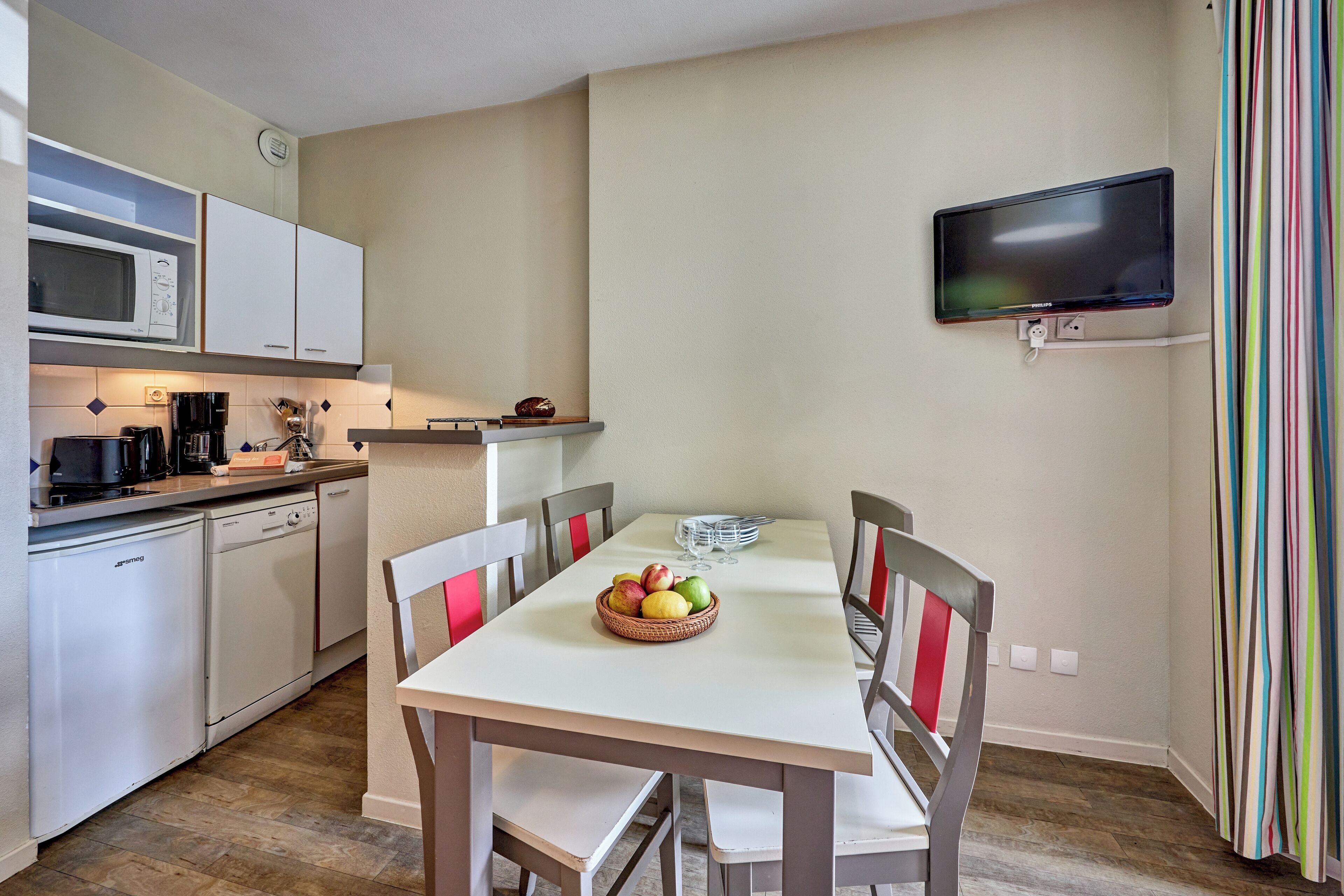 Een compacte keuken met witte apparaten, een eettafel met stoelen en een wandtelevisie.
