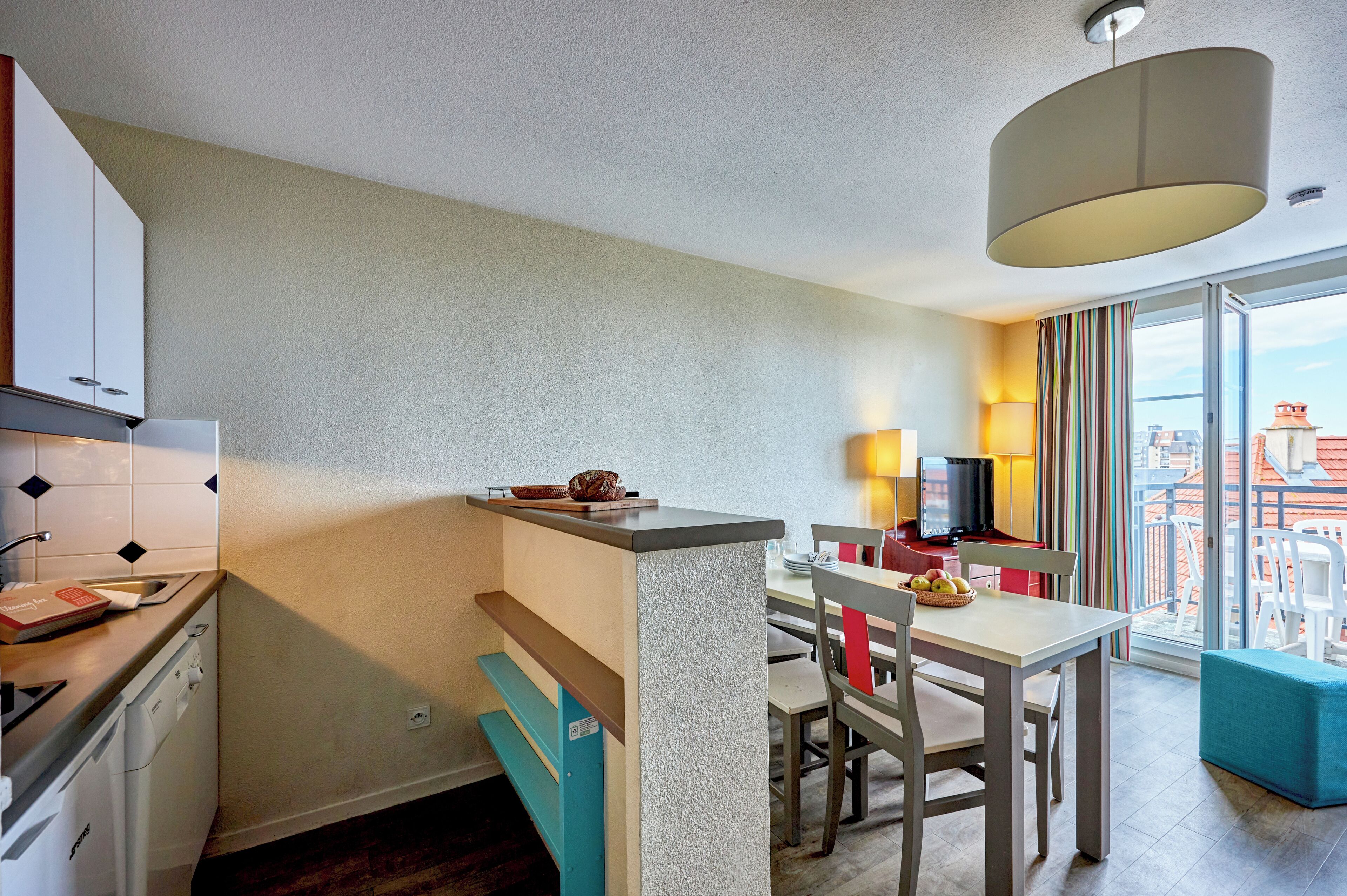 A compact kitchen with a dishwasher, a small island with a cutting board, and a dining area with a TV and a balcony with a view of the city.