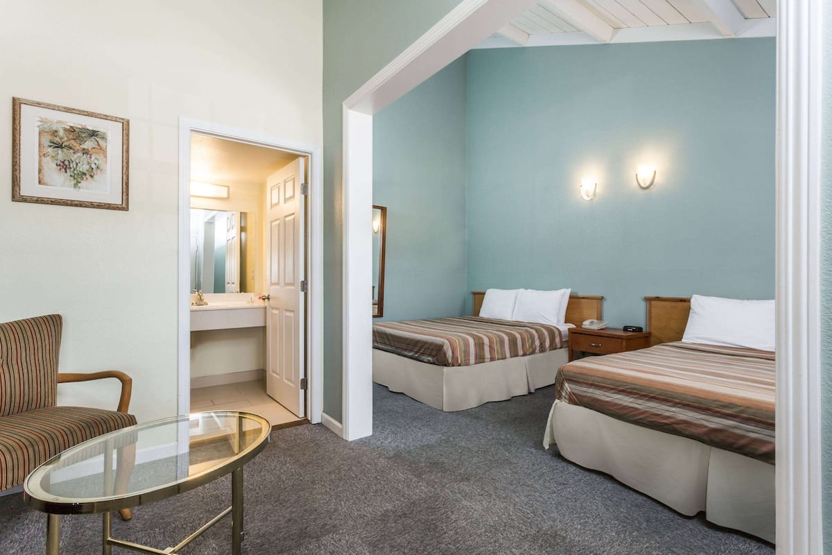 A hotel room with two beds, a sitting area with a glass table, and a bathroom visible through an open door.