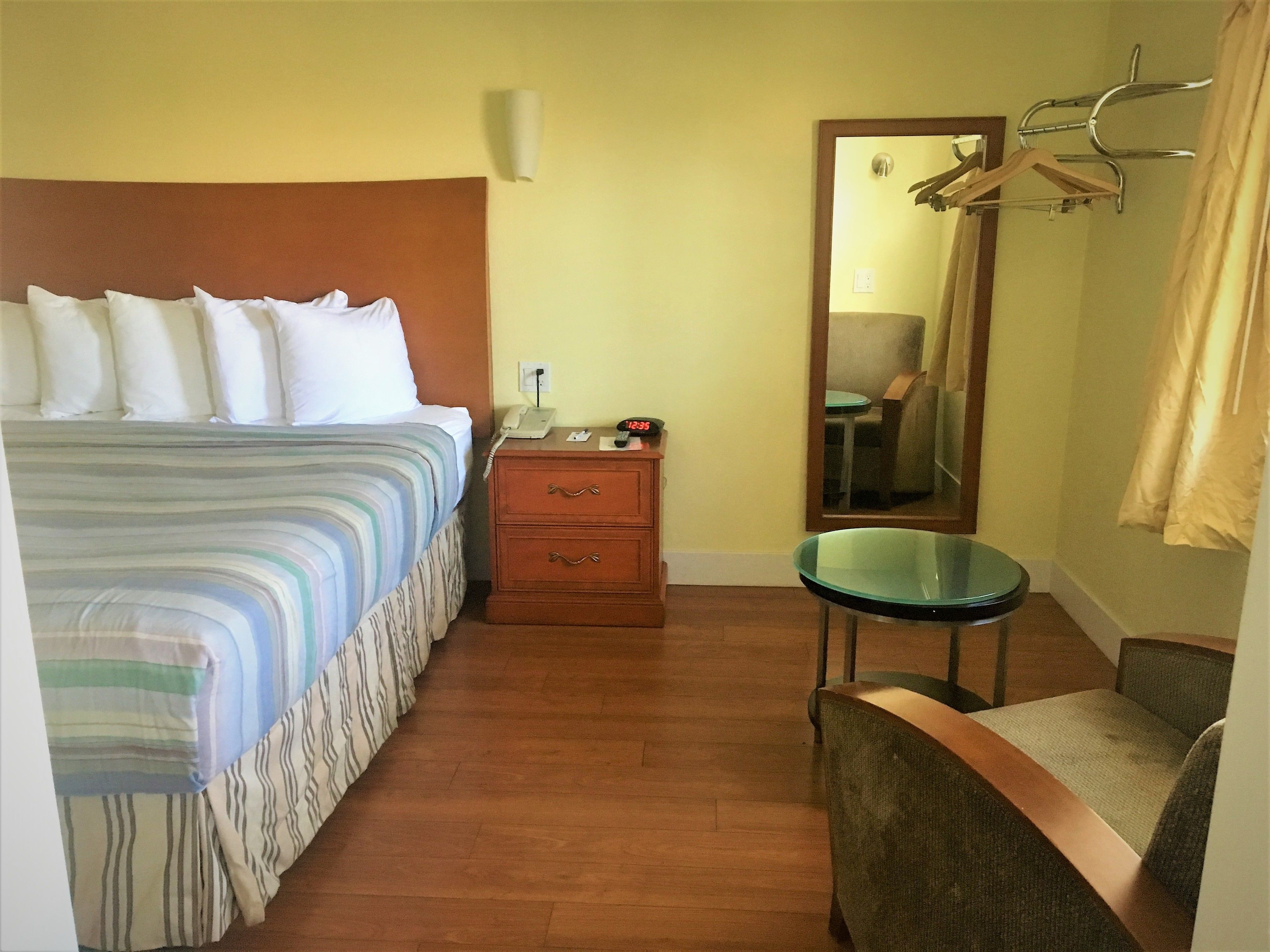 A hotel room with a bed, a bedside table with a phone, a mirror, a chair, and a coat rack.