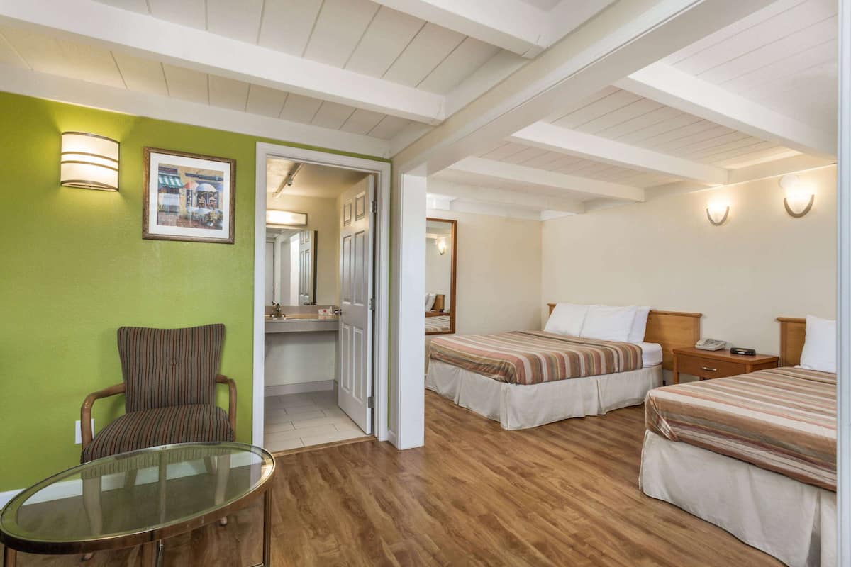 A hotel room with two beds, a chair, a glass table, and a bathroom visible through an open door.