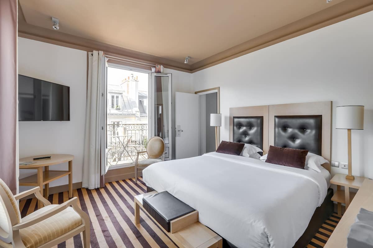 Habitación de hotel con una cama grande, un escritorio, una silla y un balcón con vistas a edificios.
