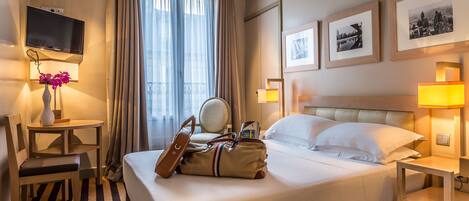 Ein Hotelzimmer mit Bett, Nachttischen, Fernseher, Schreibtisch mit Stuhl, Lampe, Blumen in einer Vase und gerahmten Bildern an der Wand.
