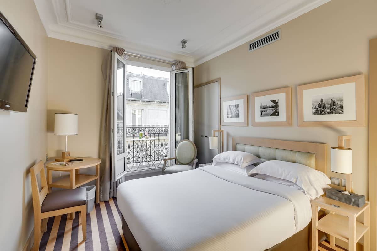 Habitación de hotel con una cama grande, un escritorio con una silla, una mesita, un televisor y un balcón con vistas a edificios.