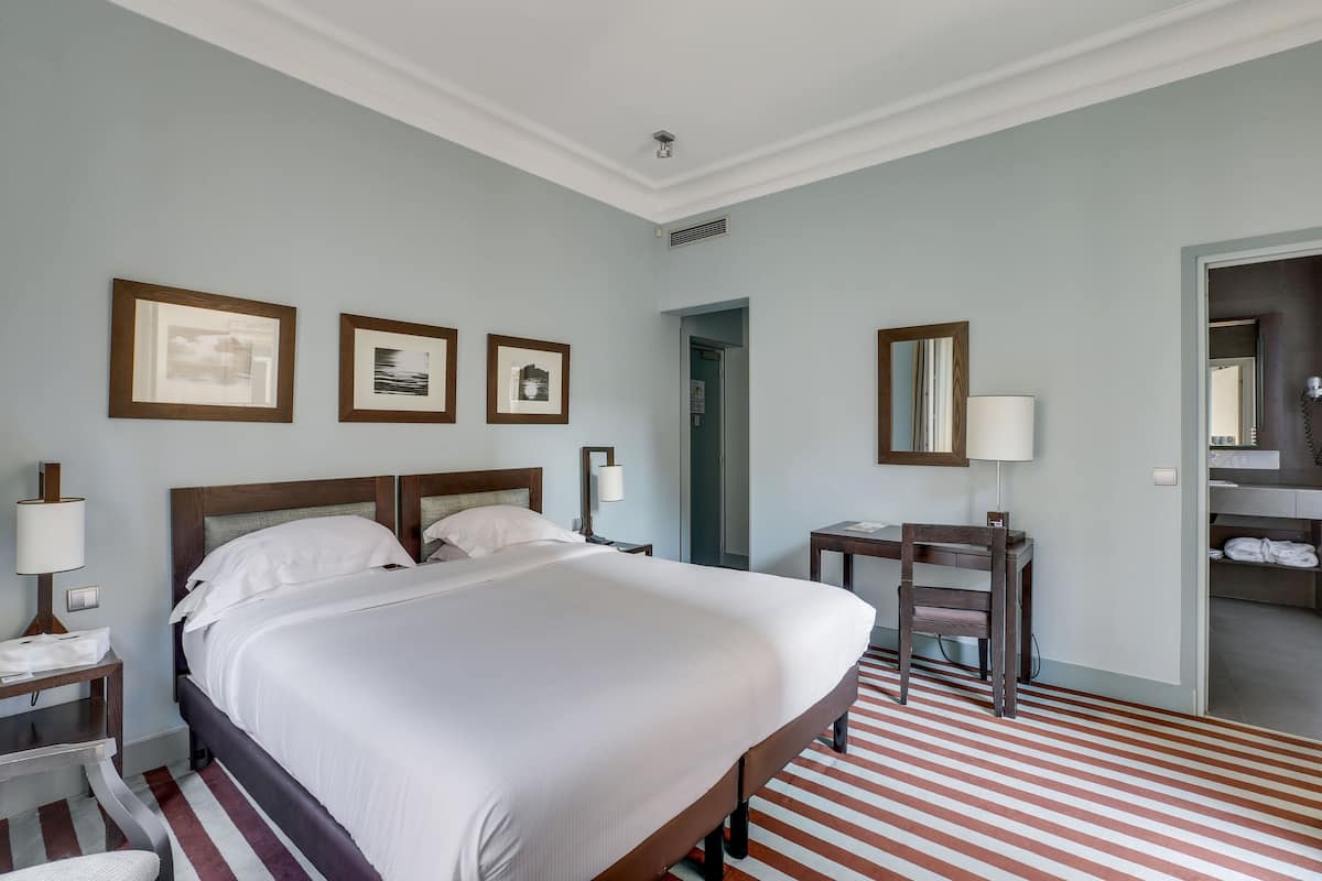 Una habitación de hotel con una cama grande, un escritorio con silla, un espejo y cuadros en la pared.