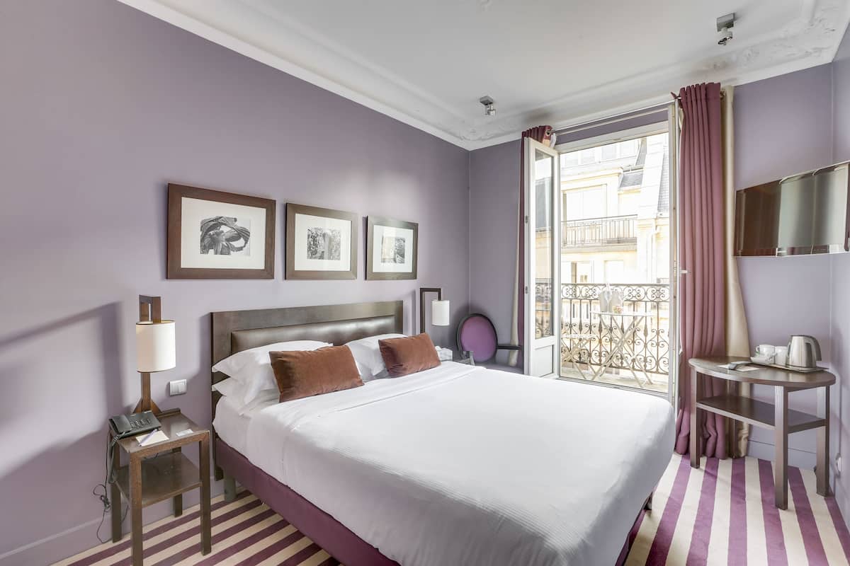 Habitación de hotel con cama, mesita de noche con lámpara, una pequeña mesa con una taza y un hervidor, y un balcón con vistas.