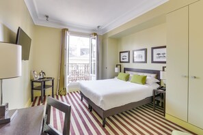 A hotel room with a bed, a desk, a chair, a TV, and a balcony with curtains. - Hotel Duret (Paris)