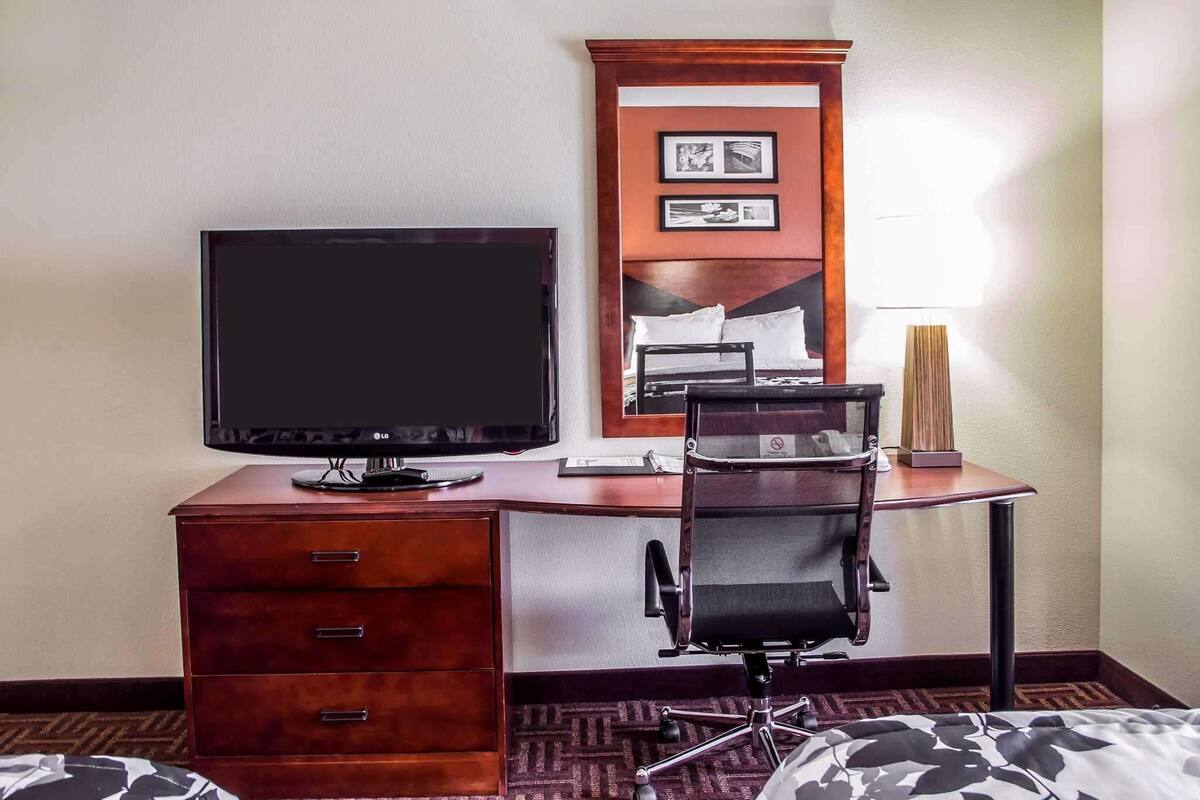 Ein Hotelzimmer mit einem Holztisch, einem schwarzen Bürostuhl, einem Flachbildfernseher, einem Bett mit gemustertem Überwurf und einem wandmontierten Spiegel mit gerahmten Bildern.