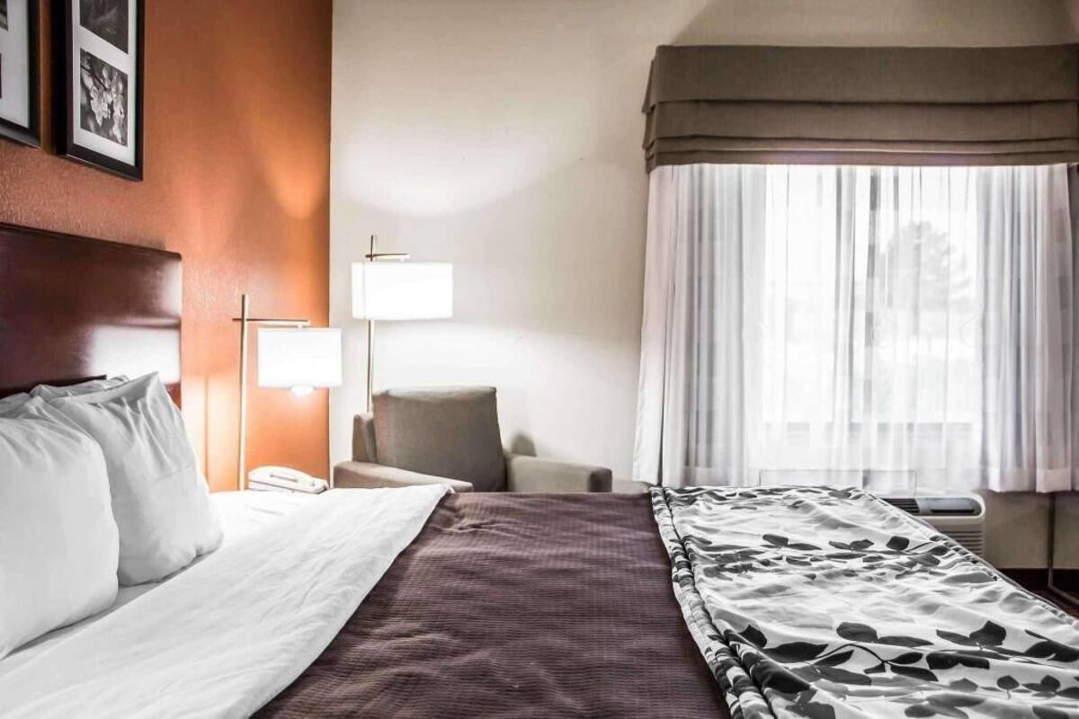 Ein Hotelzimmer mit Bett, Stuhl, Lampe, einem fenster mit Vorhängen und einem an der Wand befestigten Telefon.