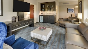 A modern living room with a flat-screen TV, a sofa, a coffee table, and a kitchen area with a refrigerator and cabinets.