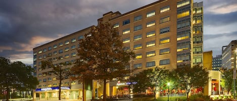 A multi-story hotel with a modern facade, multiple ground-level retail spaces, and a well-lit entrance.
