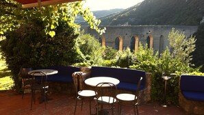 An outdoor patio with tables and chairs, a red canopy, and a view of a mountain and a stone wall with arches. - Hotel Gattapone (Spoleto)