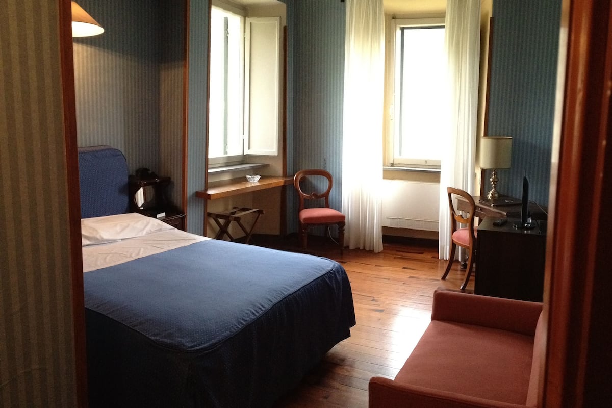 Quarto de hotel com cama, escrivaninha, duas cadeiras, abajur e janela com cortinas.