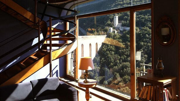 A room with a spiral staircase, a large window with a view of a forest and a tower, a table with a lamp, and a bookshelf. - Hotel Gattapone (Spoleto)