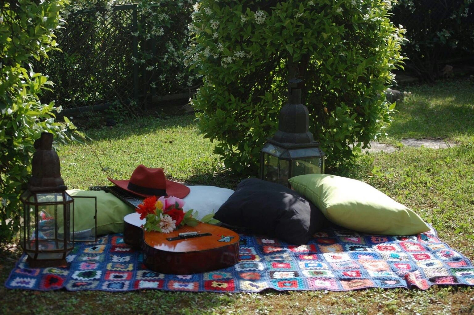 A picnic setup with a blanket, guitar, hat, lanterns, and pillows in a garden.