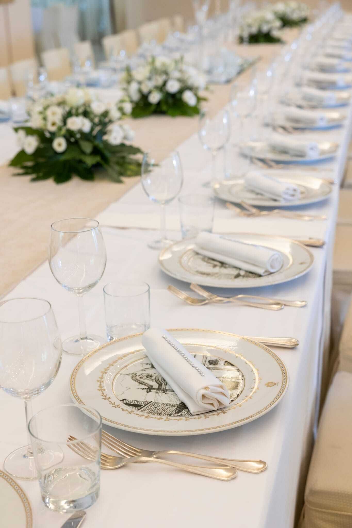 A long dining table set with white tablecloths, gold-rimmed plates, and clear glassware.