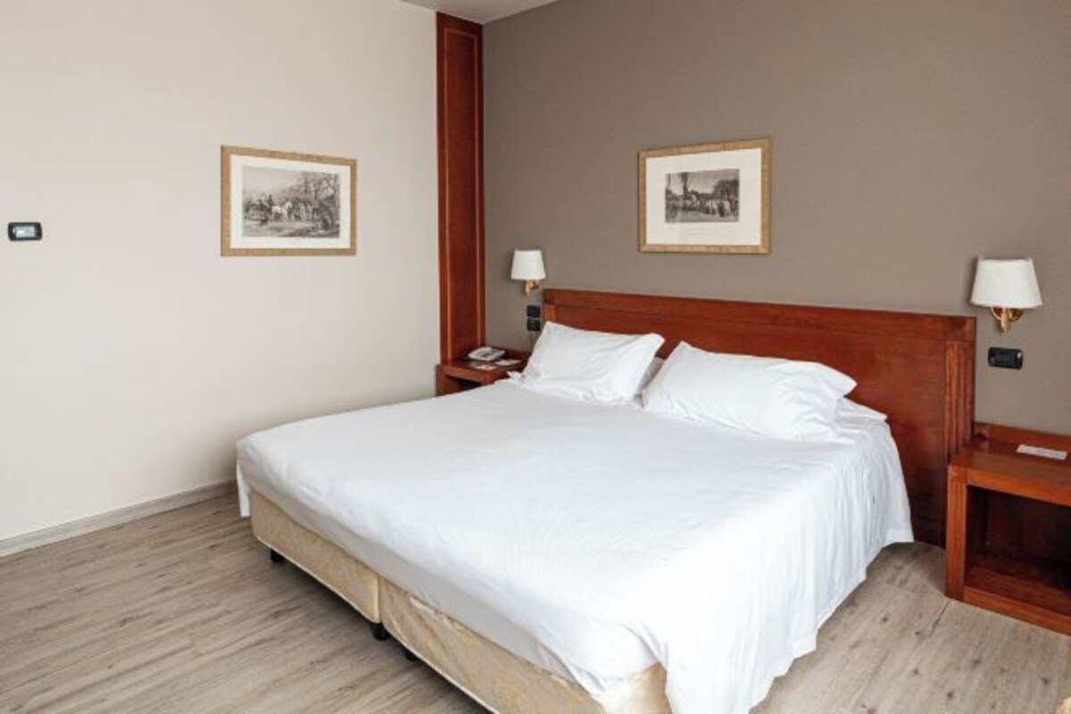 A neatly made bed in a hotel room with a wooden headboard, two bedside tables with lamps, and two framed pictures on the walls.