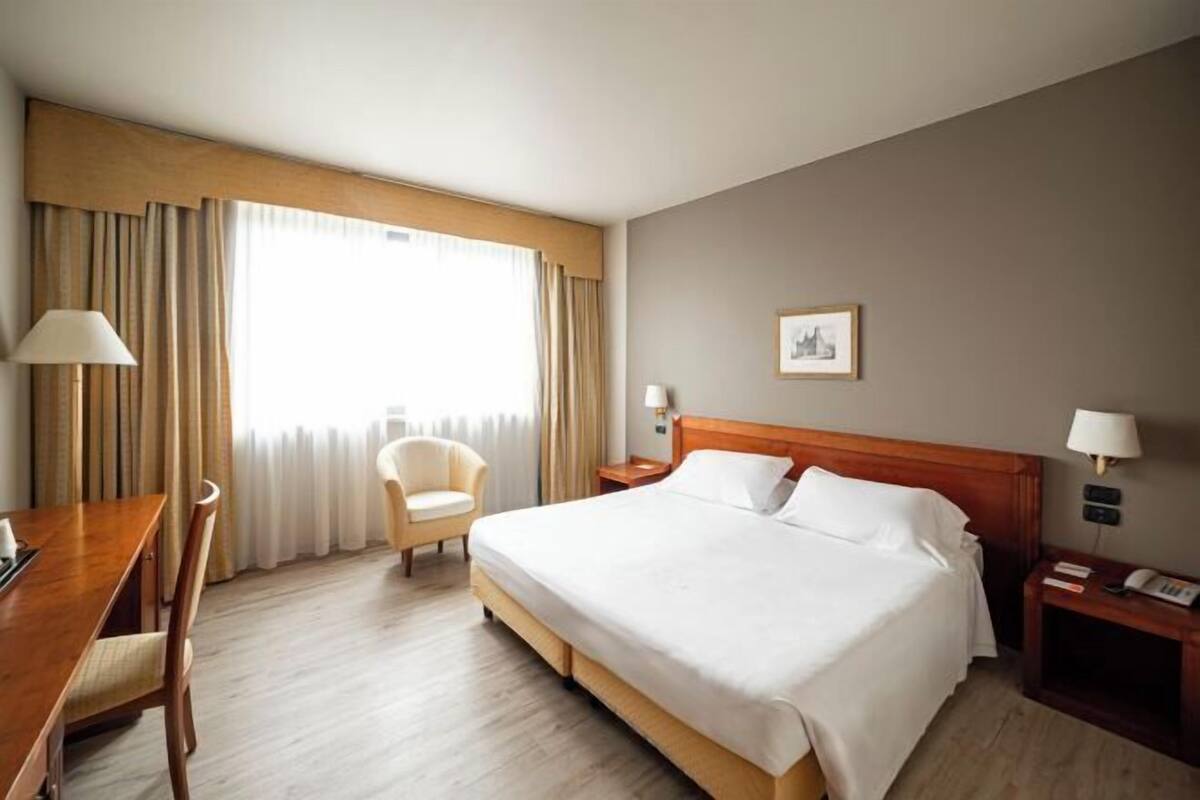 Une chambre d’hôtel comprenant un lit, un bureau, une chaise, une table de chevet, une lampe et une fenêtre avec des rideaux.
