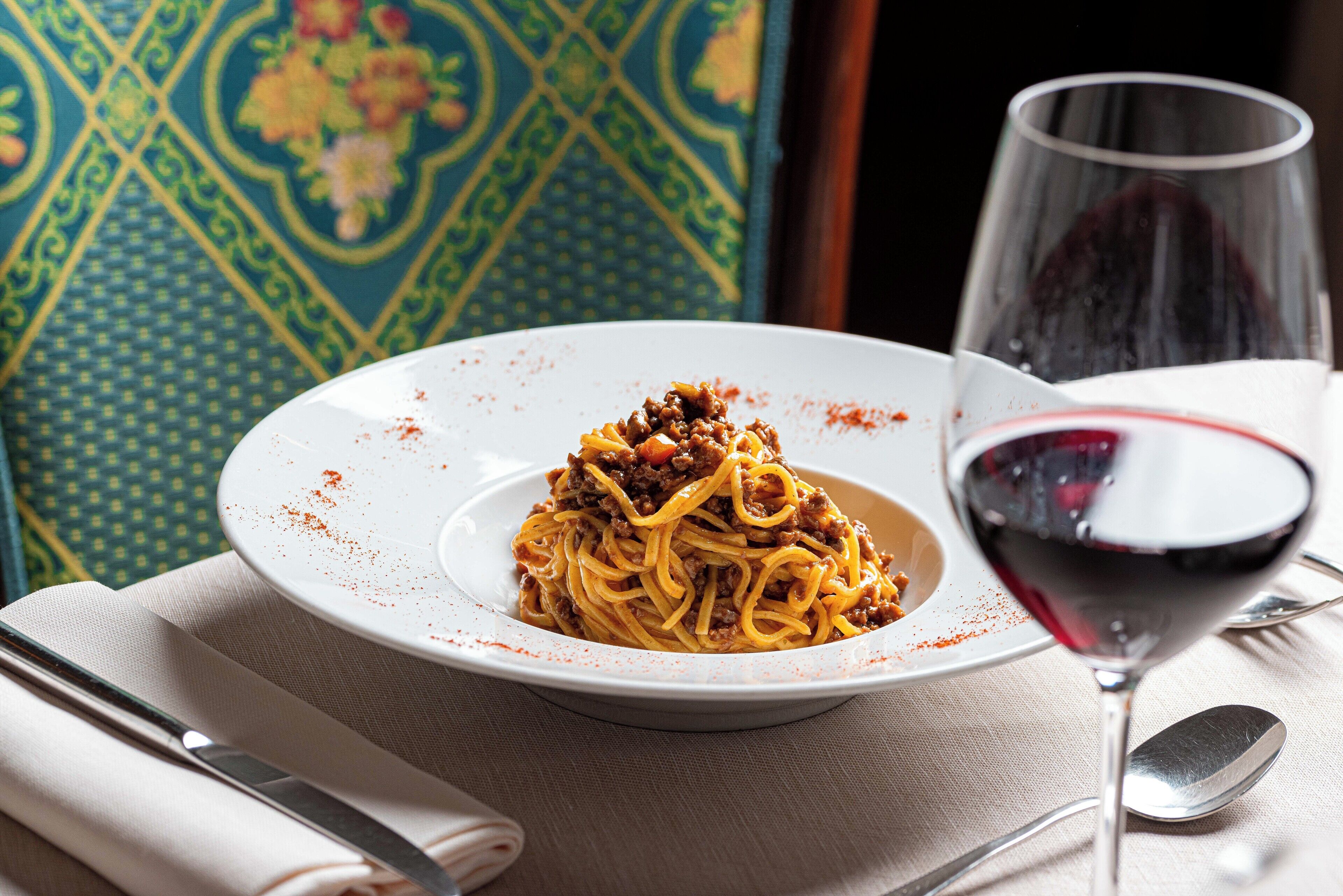 A plate of spaghetti with meat sauce, a glass of red wine, and a spoon on a table with a napkin and a knife.