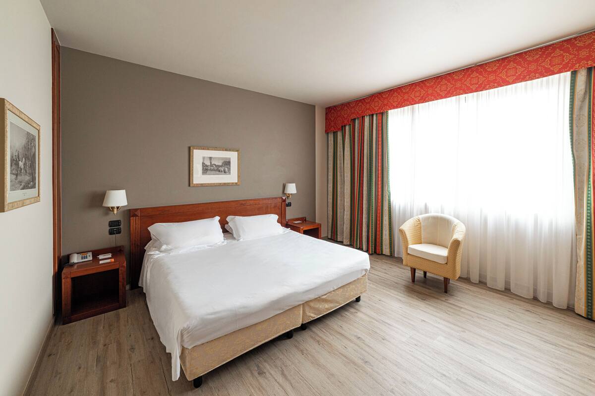 Une chambre d’hôtel avec un grand lit, deux tables de chevet avec des lampes, une chaise, un tableau encadré au mur et une fenêtre avec des rideaux.