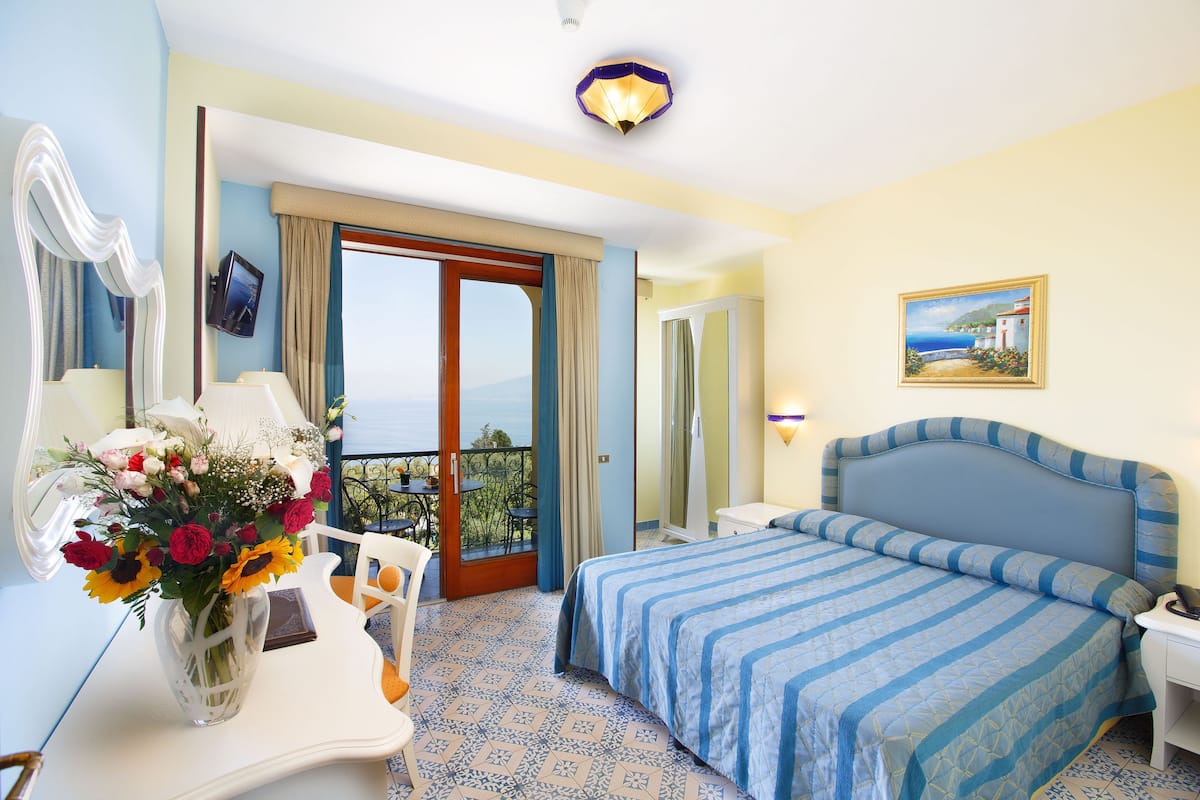 Hotelkamer met een bed, een bureau met een bloevas, een schilderij aan de muur en een balkon met uitzicht.