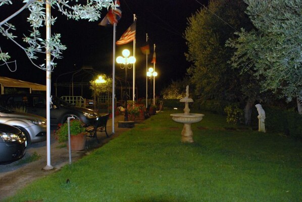 Un espace extérieur nocturne avec une voiture garée à gauche, une fontaine au centre et une statue à droite.
