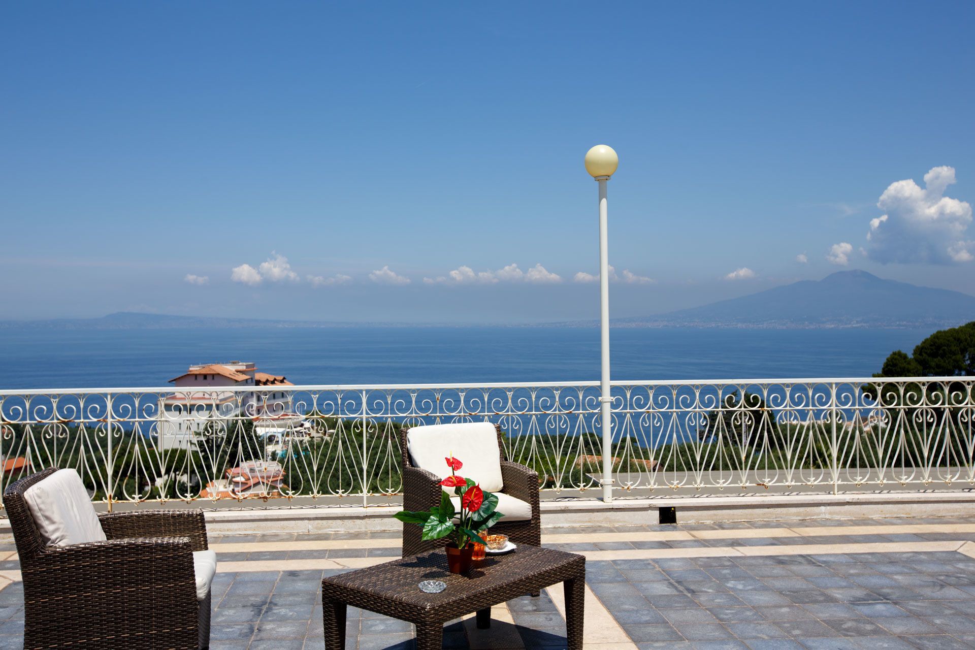 Un balcón con muebles de mimbre, una planta en maceta y vistas al mar y montañas a lo lejos.