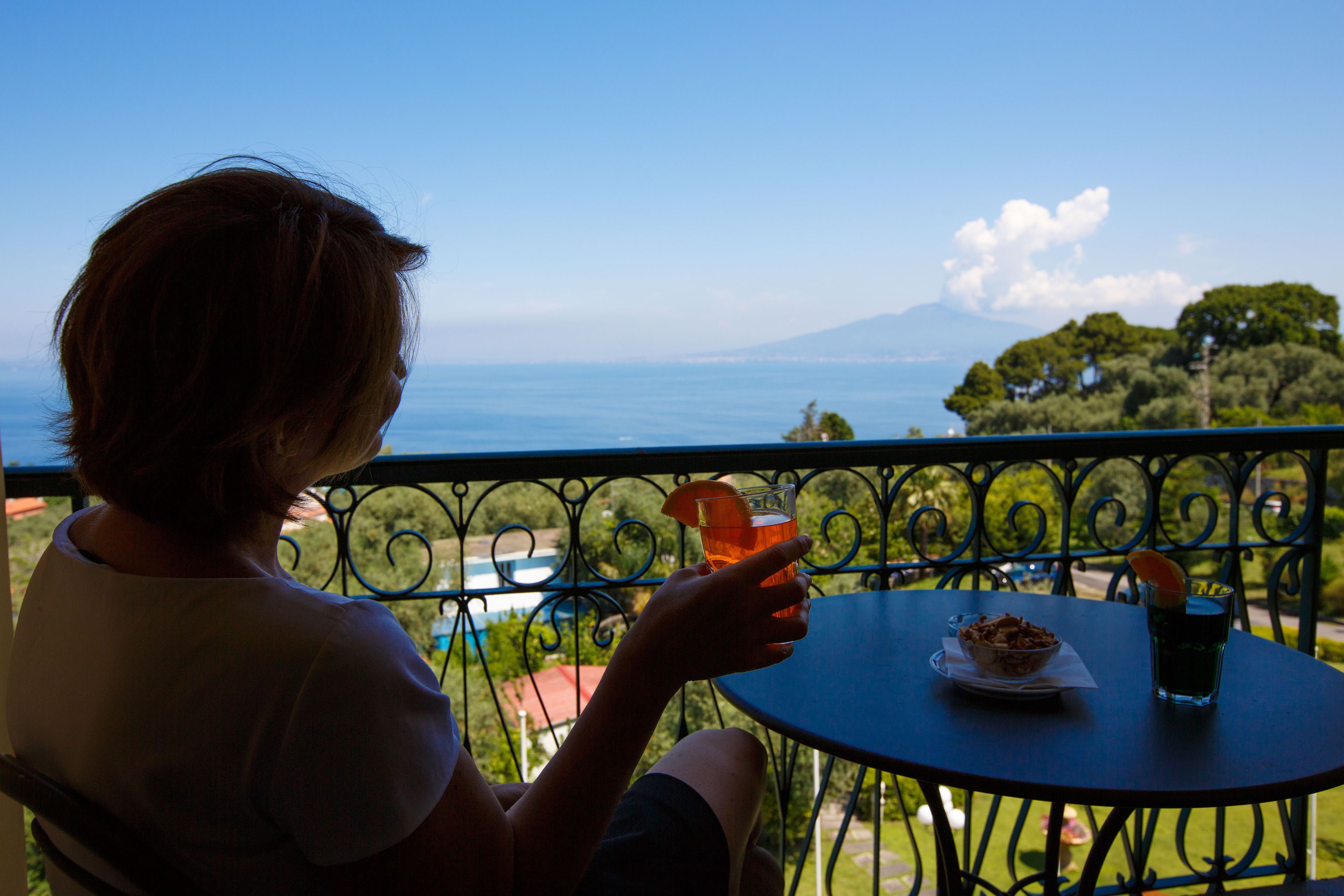 Una persona sentada a una mesa con una bebida y un plato de comida, con vista a un paisaje marítimo y montañoso.