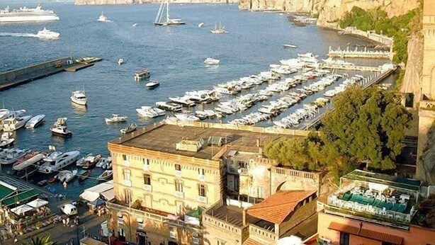 Un port de plaisance où sont amarrés de nombreux bateaux, un grand navire de croisière et un bâtiment historique doté d’une terrasse offrant une vue sur l’eau.