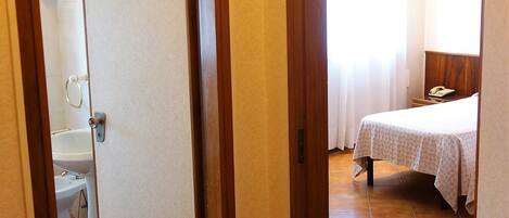Une chambre d’hôtel avec une salle de bain visible à travers une porte, un lit recouvert d’une courtepointe blanche et une table de chevet avec une lampe.