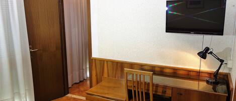 A small room with a wooden desk, a chair, a television mounted on the wall, and a door with curtains.
