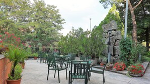 Un patio al aire libre con sillas y mesas de metal verde, rodeado de macetas con plantas y un muro de piedra.
