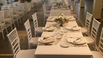 Una larga mesa de banquete dispuesta para un evento formal, rodeada de sillas blancas, con una habitación bien iluminada y un amplio ventanal al fondo.