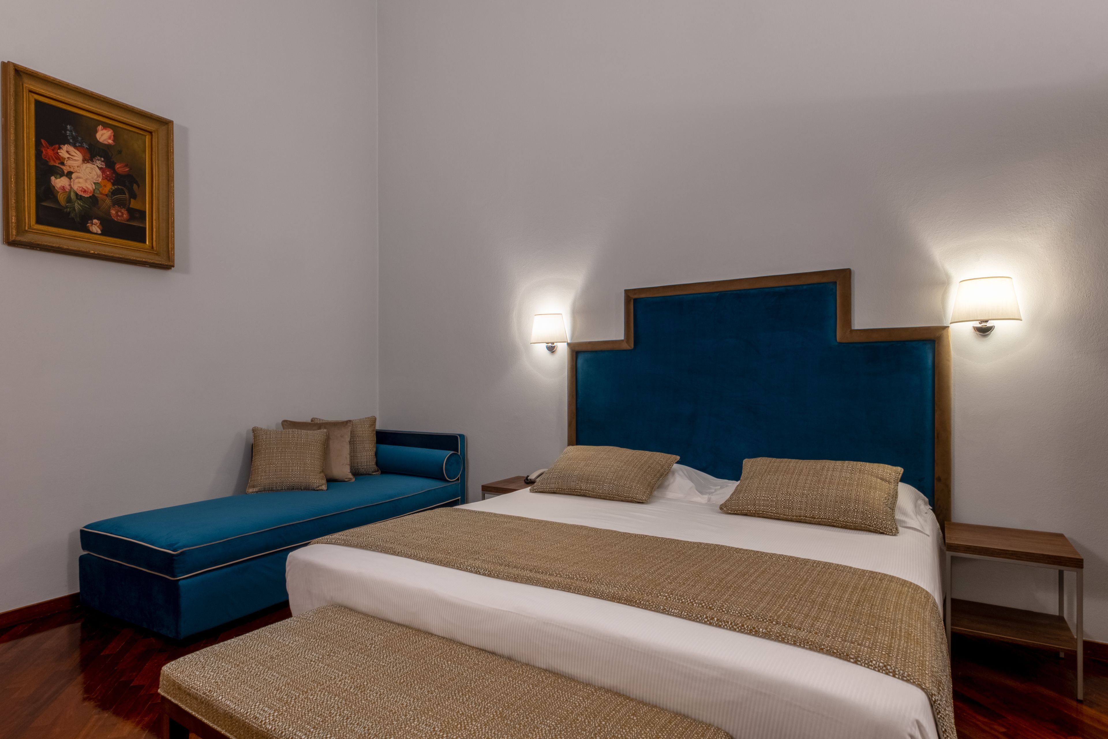 A bedroom with a large bed, a blue sofa, two bedside tables, and a wall-mounted lamp.