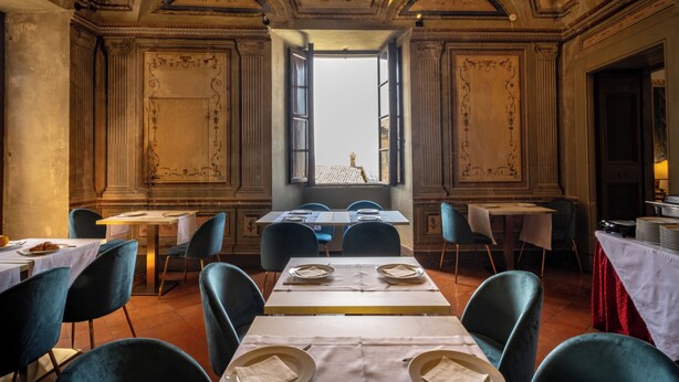 A dining area with tables set for two, blue chairs, and a view through an open window. - Hotel Bosone Palace (Gubbio)