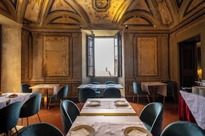 A dining area with tables set for two, blue chairs, and a view through an open window. - Hotel Bosone Palace (Gubbio)