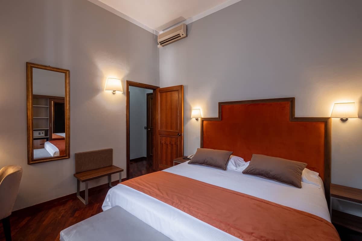 Hotellihuone, jossa on suuri sänky, kaksi yöpöytälamppua, puinen sängynpääty, penkki, peili ja ilmastointilaite.