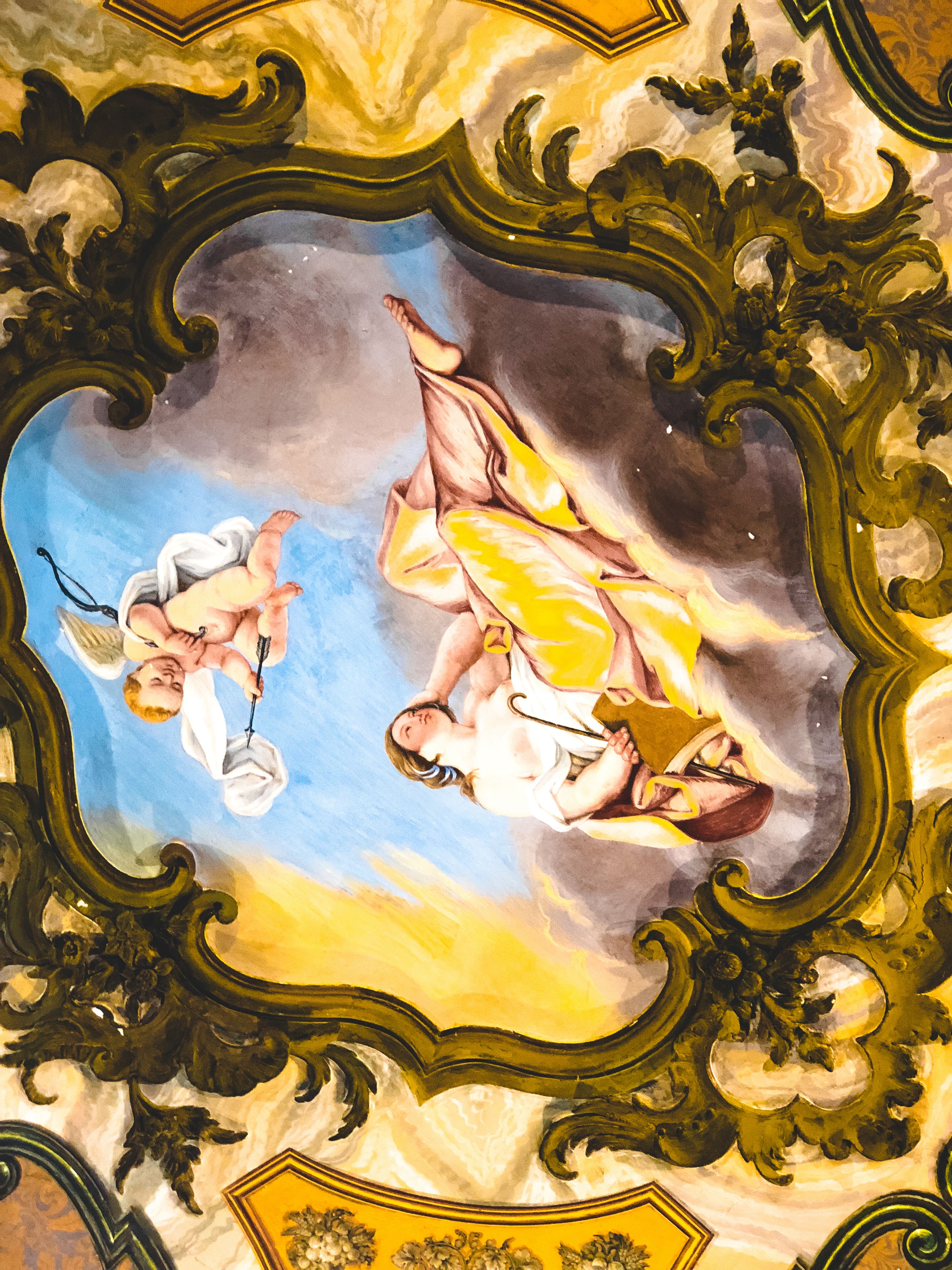 Un soffitto con elaborate decorazioni dorate e un dipinto centrale raffigurante putti.