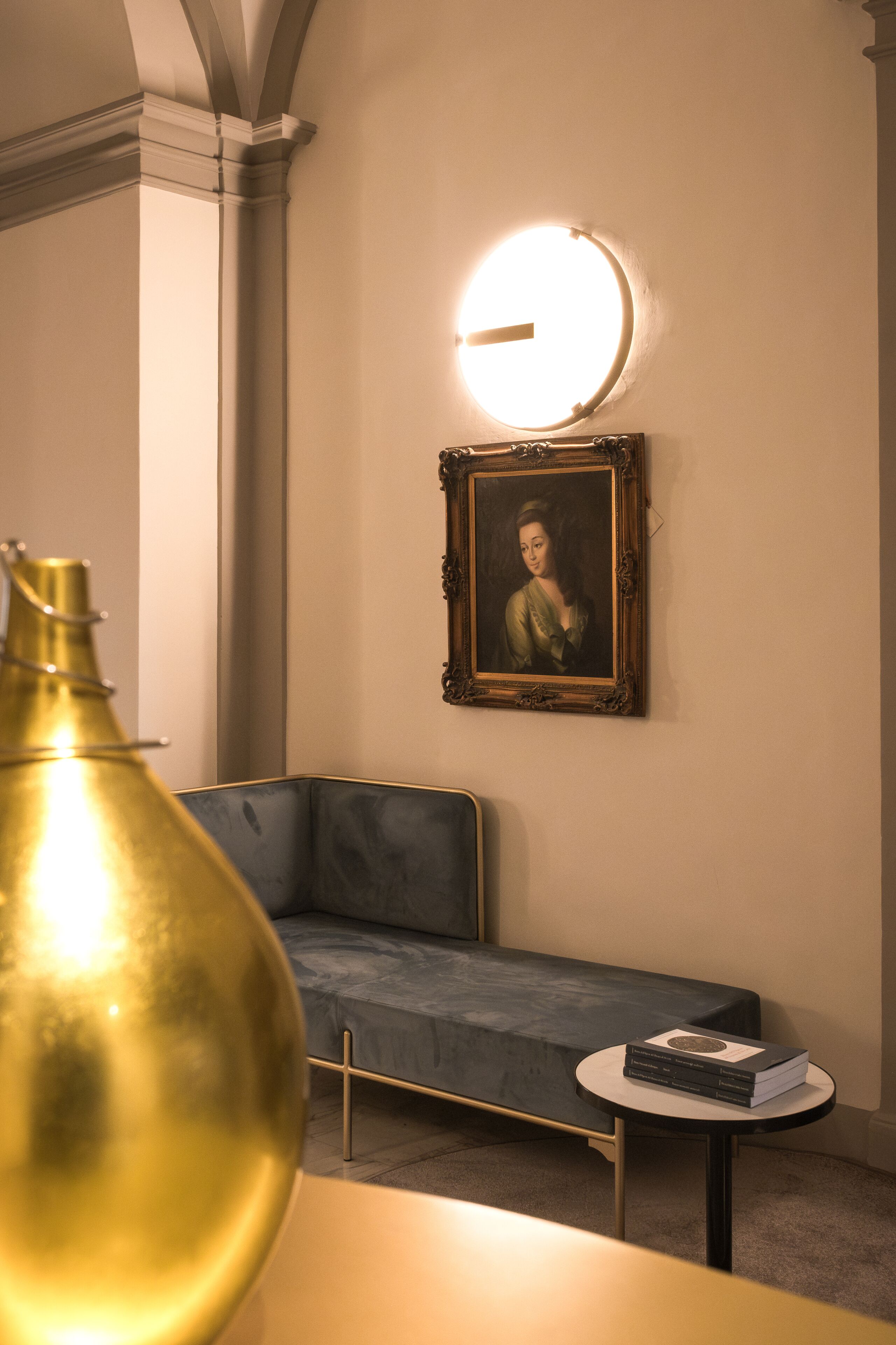 A room with a sofa, a small round table with a book, a gold-colored vase, and a framed painting on the wall.