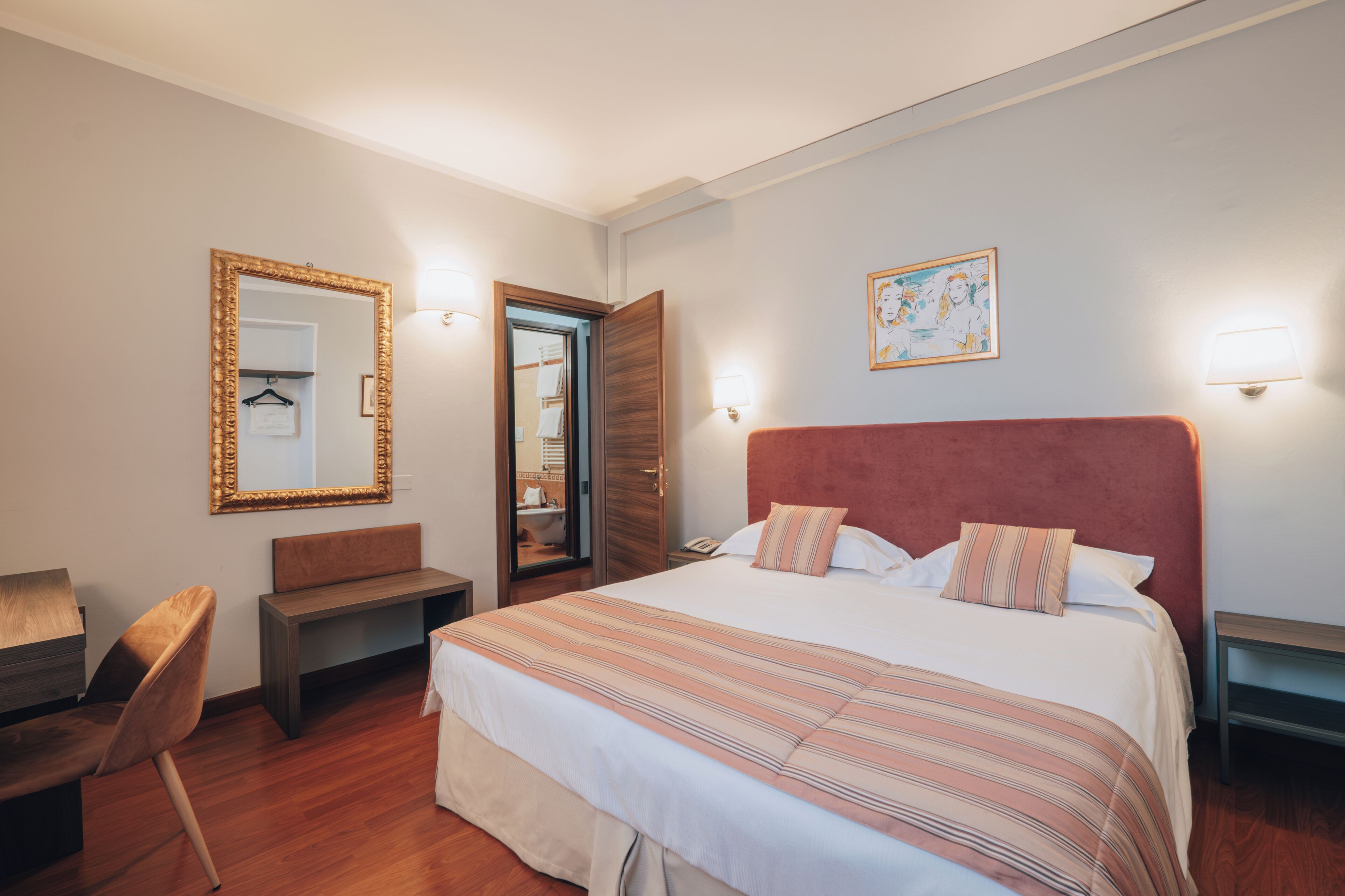 Een hotelkamer met een groot bed, een houten bankje, een bureau en een spiegel met gouden lijst.