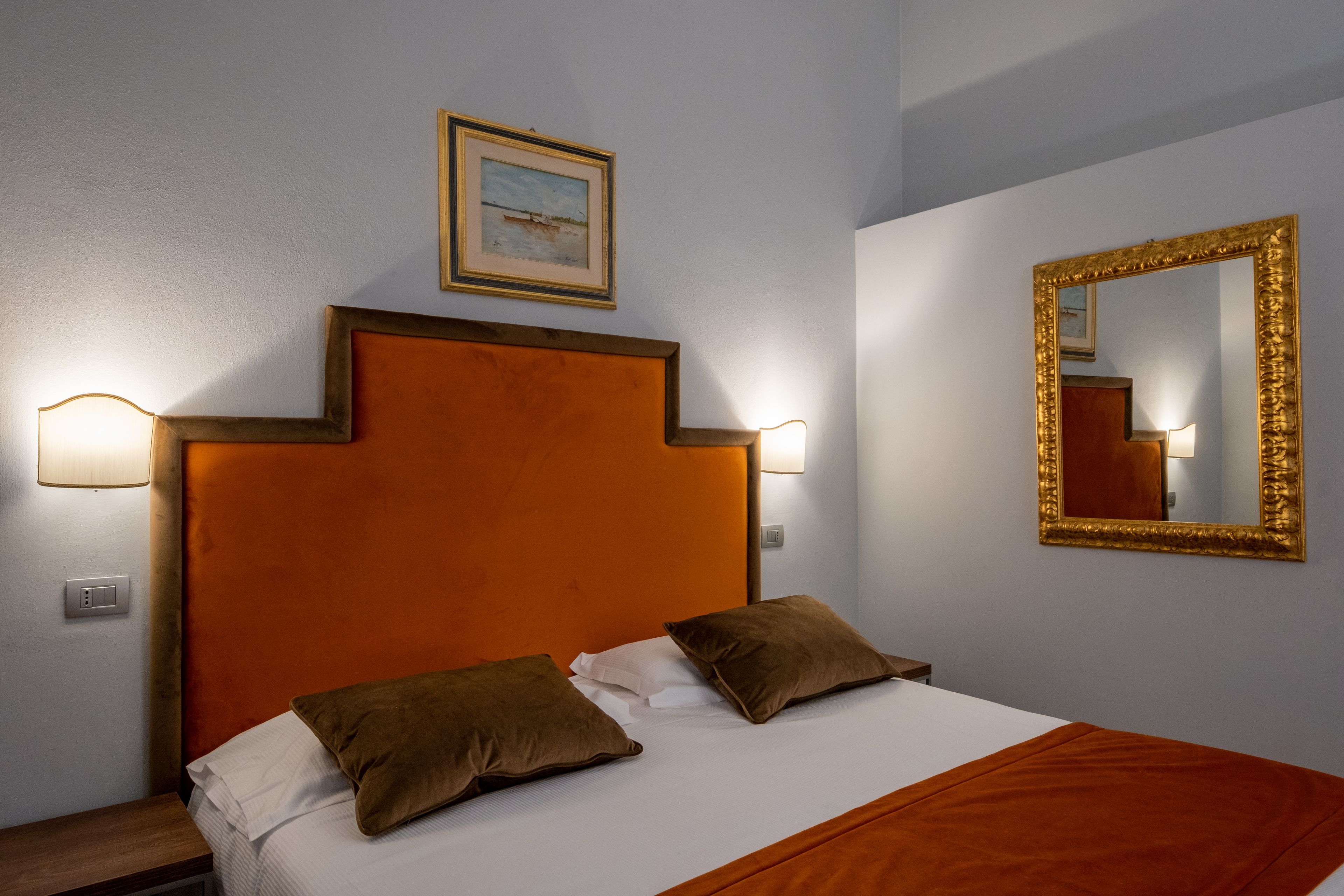 A hotel room with a bed, two pillows, a nightstand, a lamp, a framed picture on the wall, and a mirror with a gold frame.