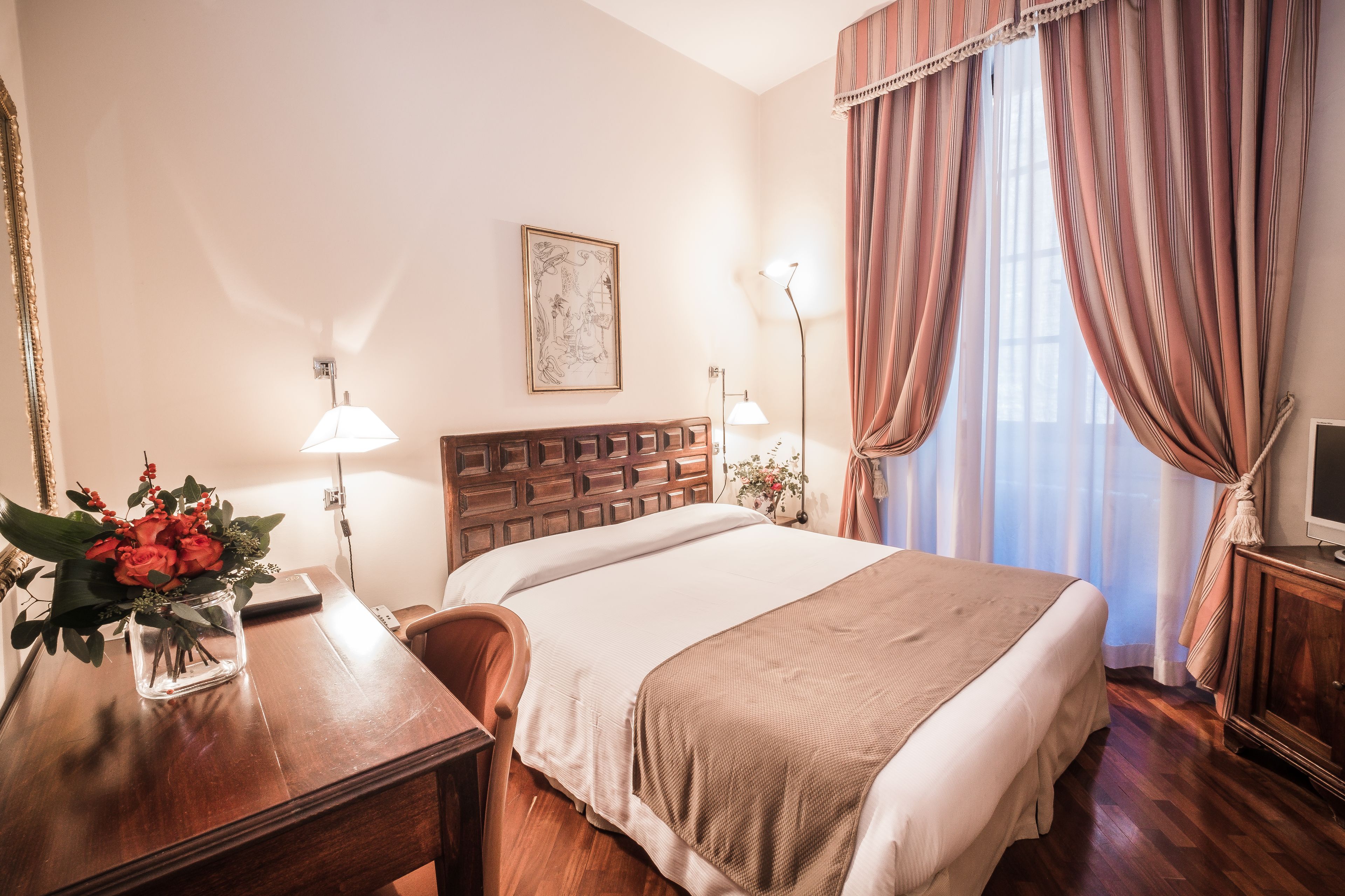 A hotel room with a wooden headboard, a bed with a beige blanket, a desk with a vase of flowers, a lamp, and a television on a wooden cabinet.