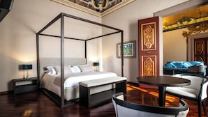 A four-poster bed with a bedside table and lamp, a round table with chairs, and a sofa in a room with ornate ceiling and wall decorations. - Hotel Bosone Palace (Gubbio)