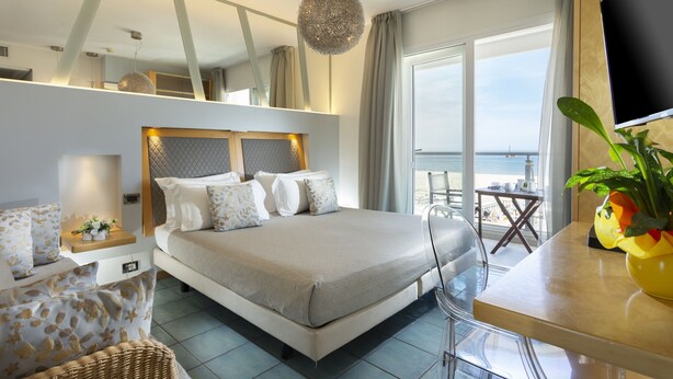 Ein modernes Hotelzimmer mit einem großen Bett, einem Essbereich mit Tisch und Stühlen, einem Fernseher und einem Balkon mit Meerblick.