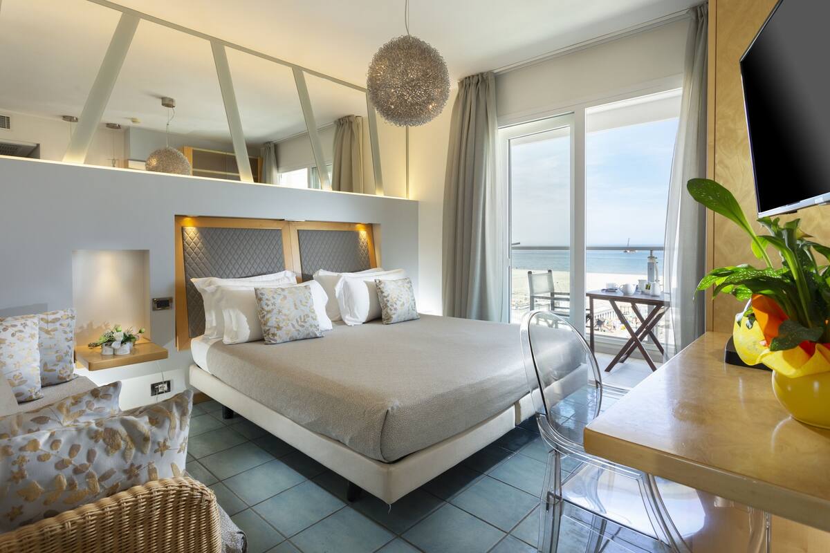 A modern hotel room with a large bed, a dining area with a table and chairs, a TV, and a balcony with a view of the sea.