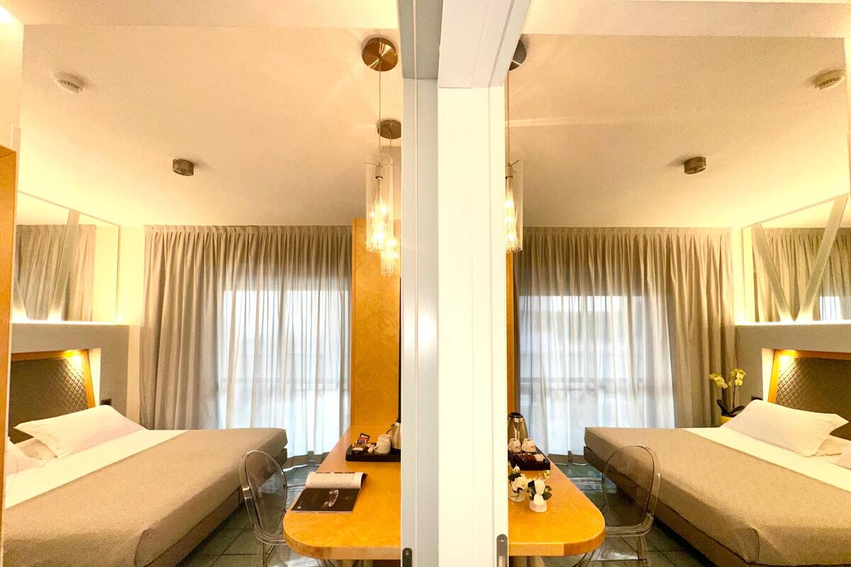 A hotel room with a bed, a desk, and a chair. There is a window with curtains and a mirror above the bed.