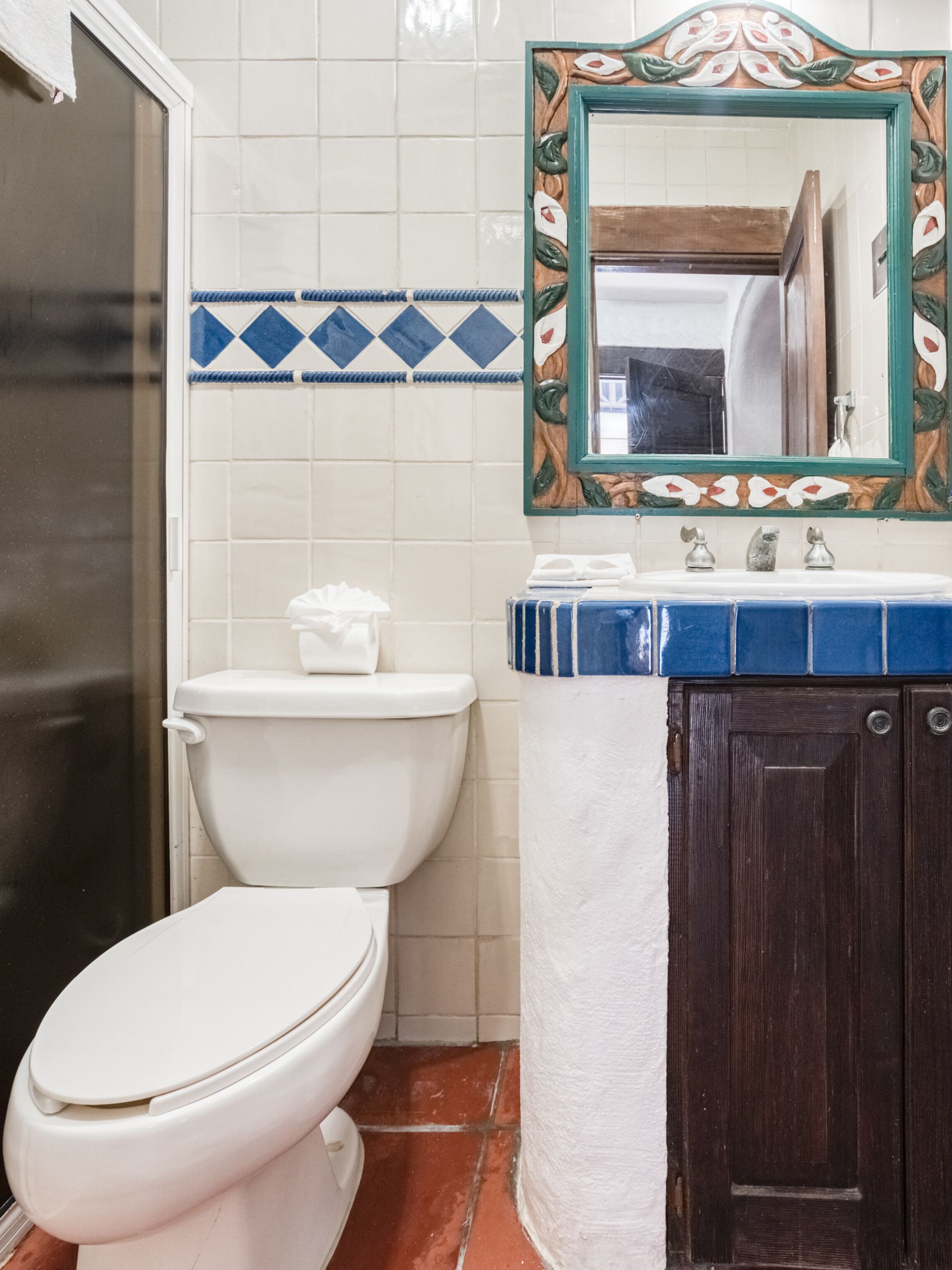 Une salle de bain avec des toilettes blanches, un lavabo doté d’un plan-vasque carrelé de bleu et de blanc, et un meuble en bois.