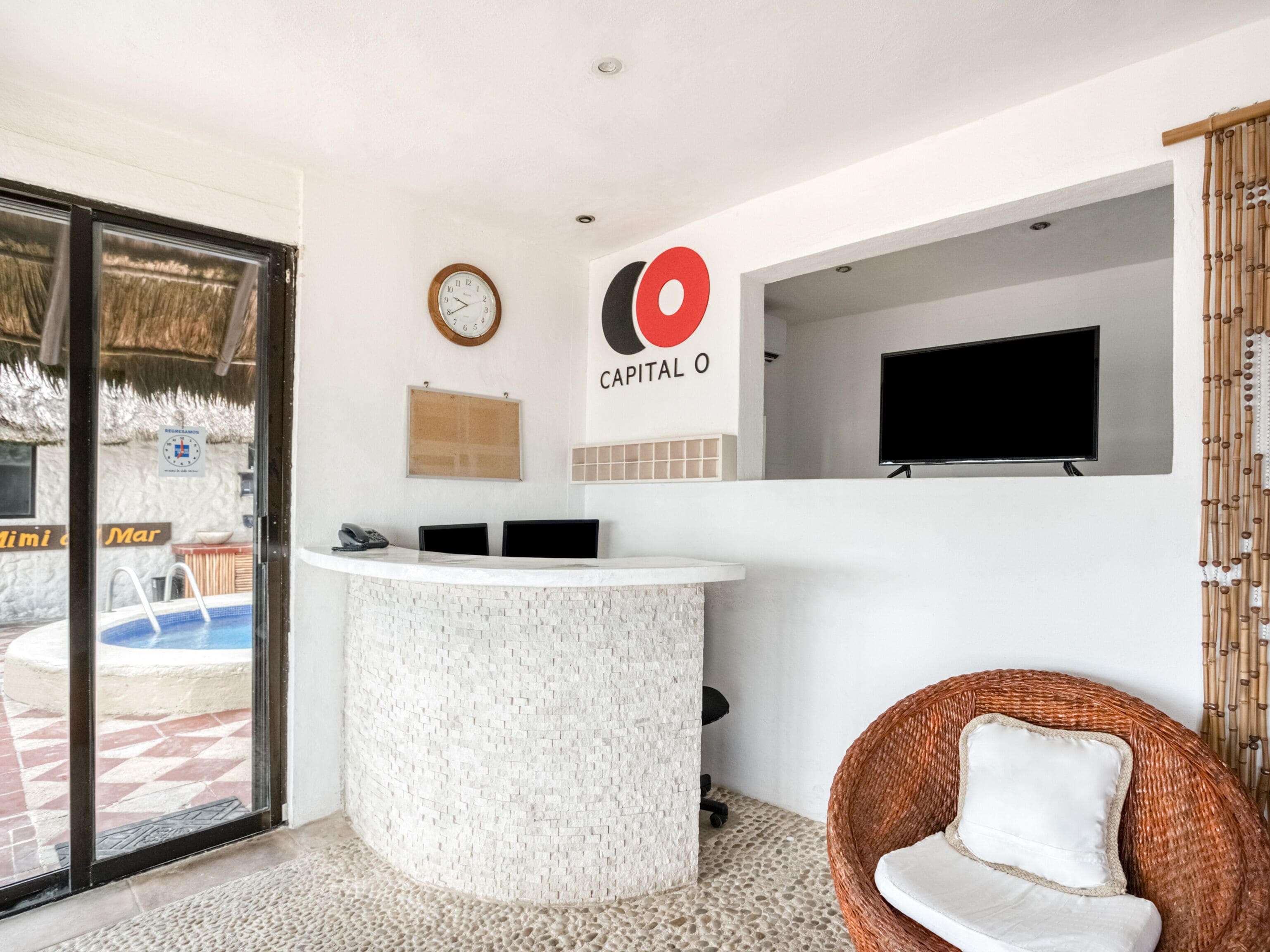 Zona de recepción con un mostrador blanco, un reloj, un televisor montado en la pared y una silla de mimbre.