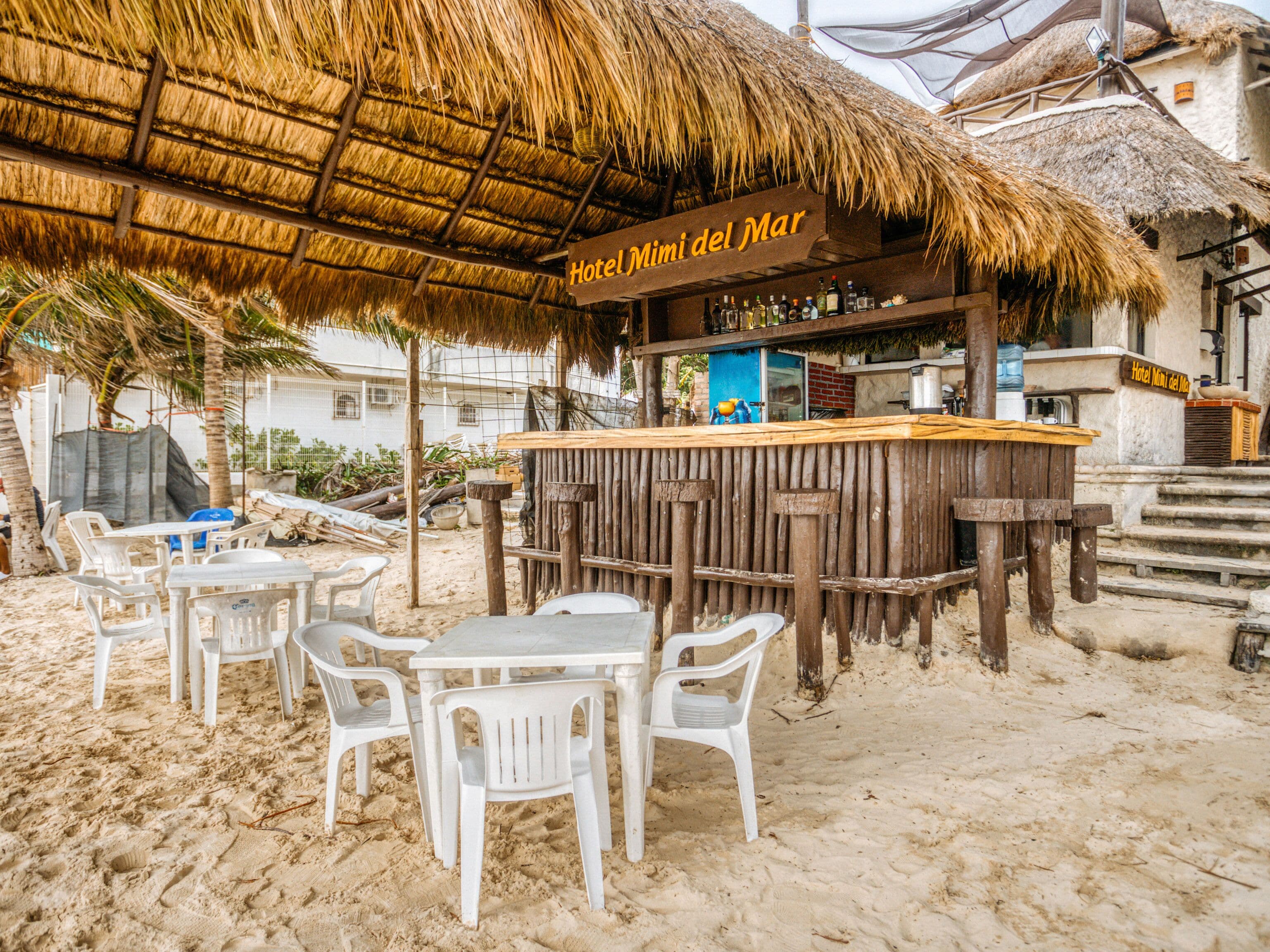 A beachside bar area with a thatched roof, white plastic chairs, and a wooden counter with a sign reading "Hotel Mini del Mar".